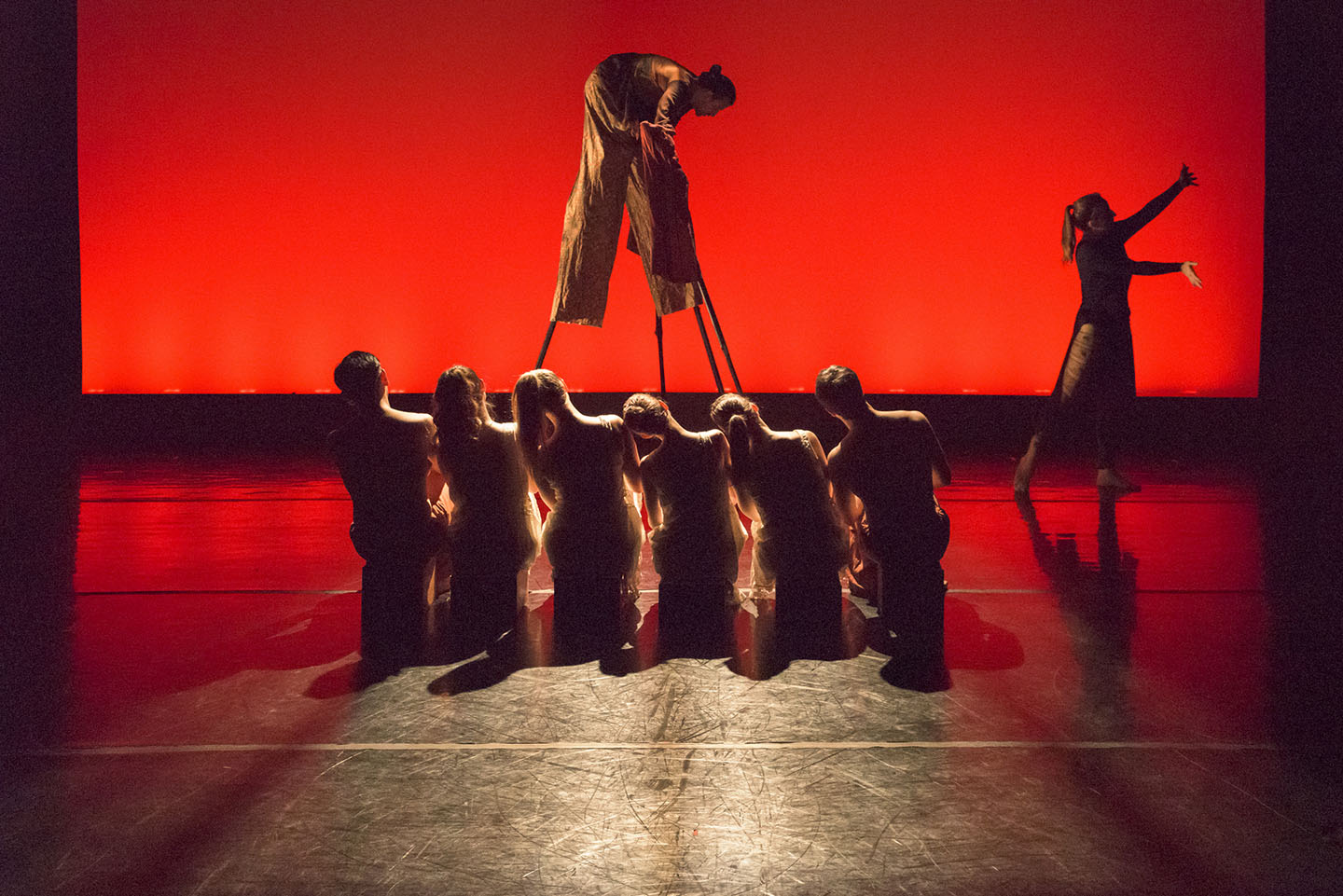 Image of 6 MorrisonDancers kneeling on a stage in front of a stilt walker in front of a red backgreound